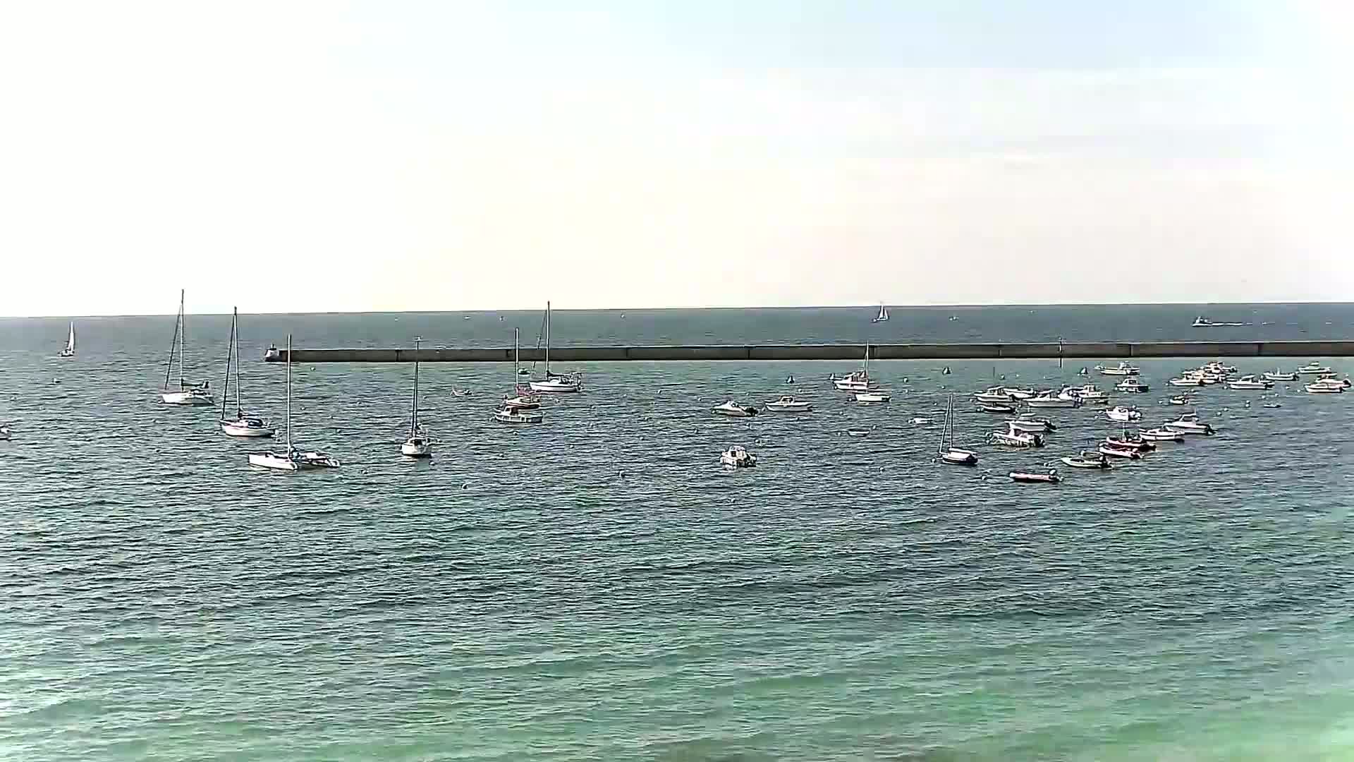 Numerous sailboats and motorboats are moored in calm, greenish-blue water behind a long, low seawall on a bright, mostly clear day.
