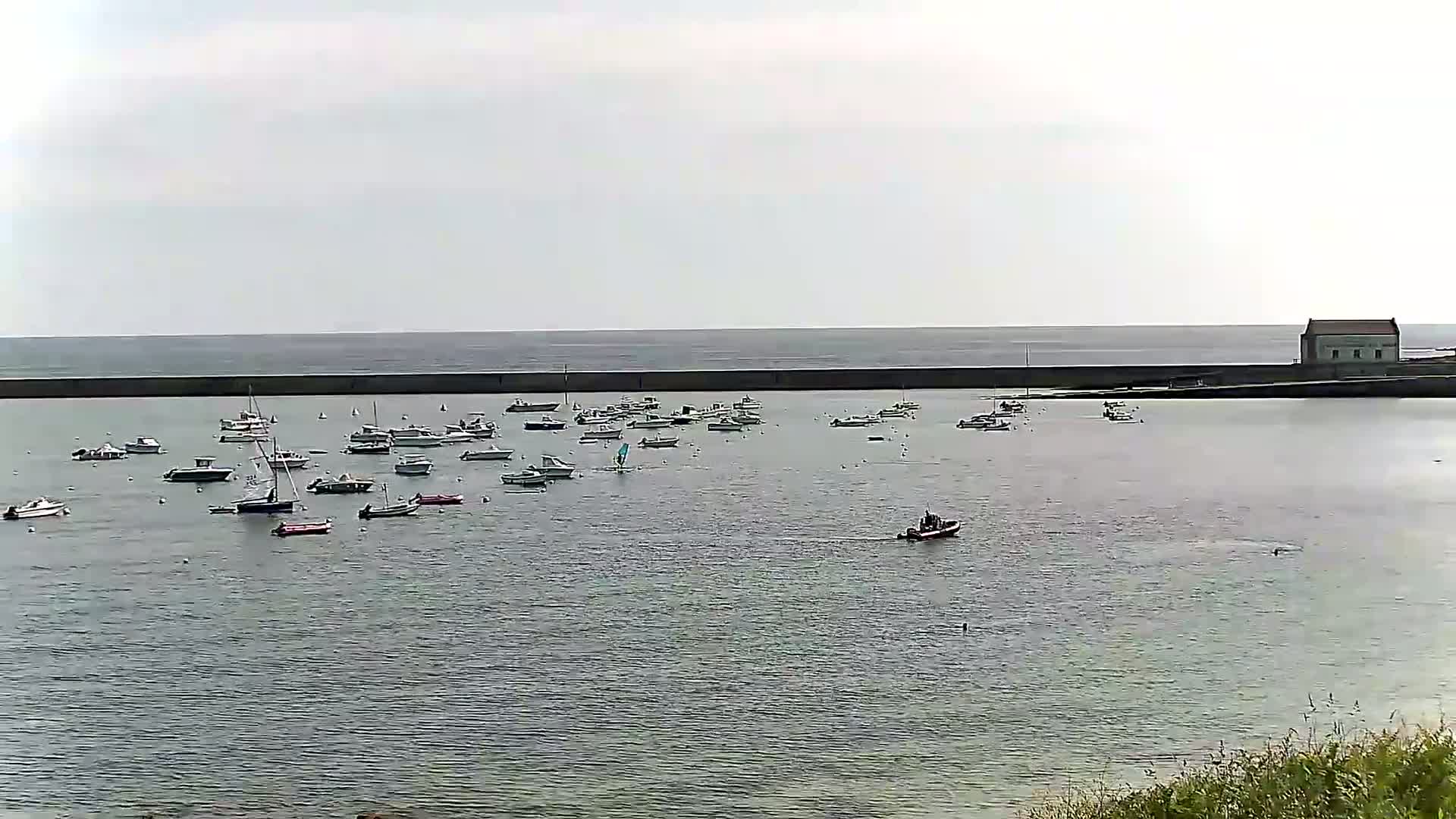 Many small boats are moored in a calm harbor protected by a seawall, with a building visible on the far right and a mostly overcast sky above.