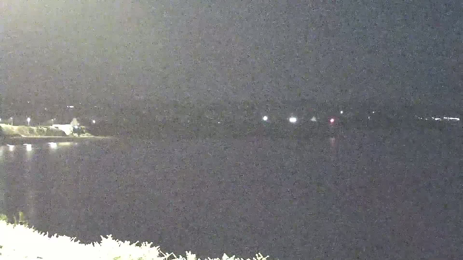 A nighttime view shows a dark body of water reflecting a few distant lights, with land visible on the far side and vegetation in the foreground.