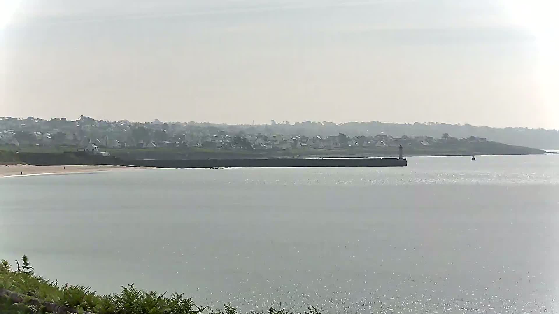 A hazy coastal scene shows a calm body of water with a distant town, a breakwater, and a small lighthouse.