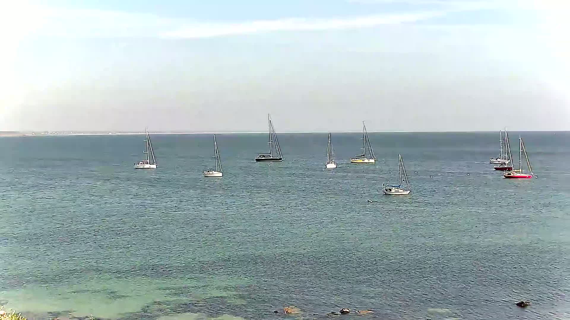 Several sailboats are anchored in calm, clear water under a mostly clear sky.
