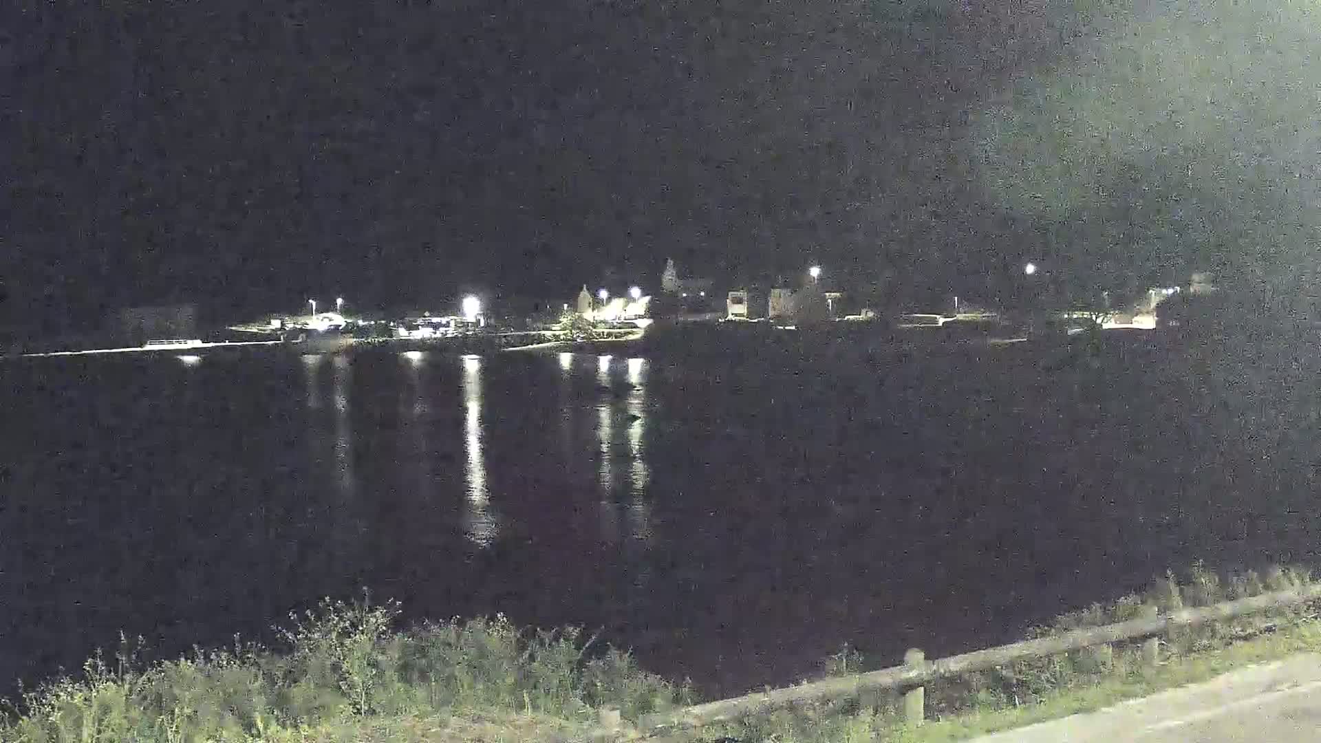A nighttime view shows a body of water reflecting lights from a town across the water, with vegetation visible in the foreground.