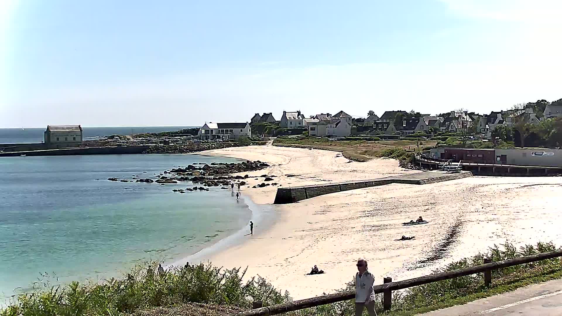 A sandy beach curves around a calm, light-blue ocean, with a small village and a few people visible in the background on a sunny day.