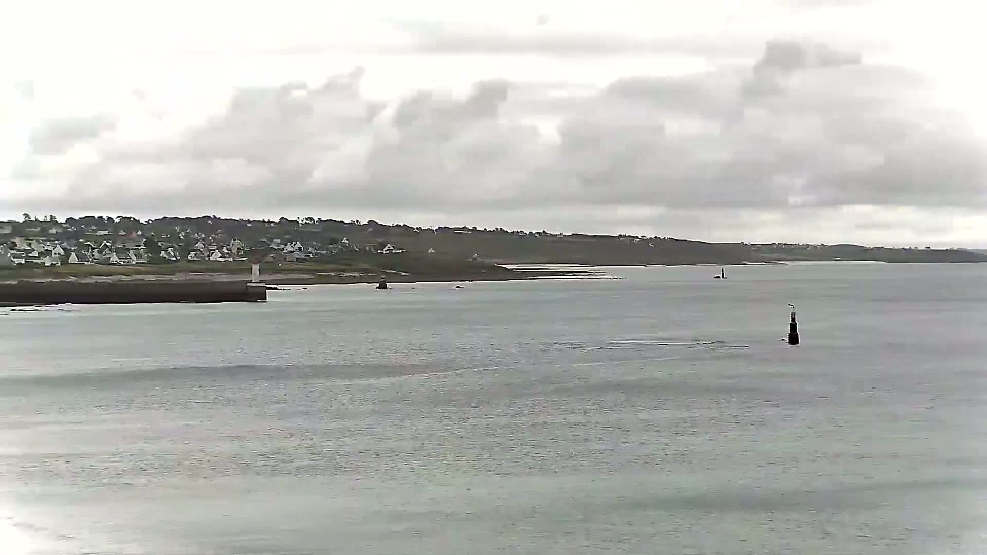 A coastal town sits on a hill overlooking a calm, grey sea under a cloudy sky.