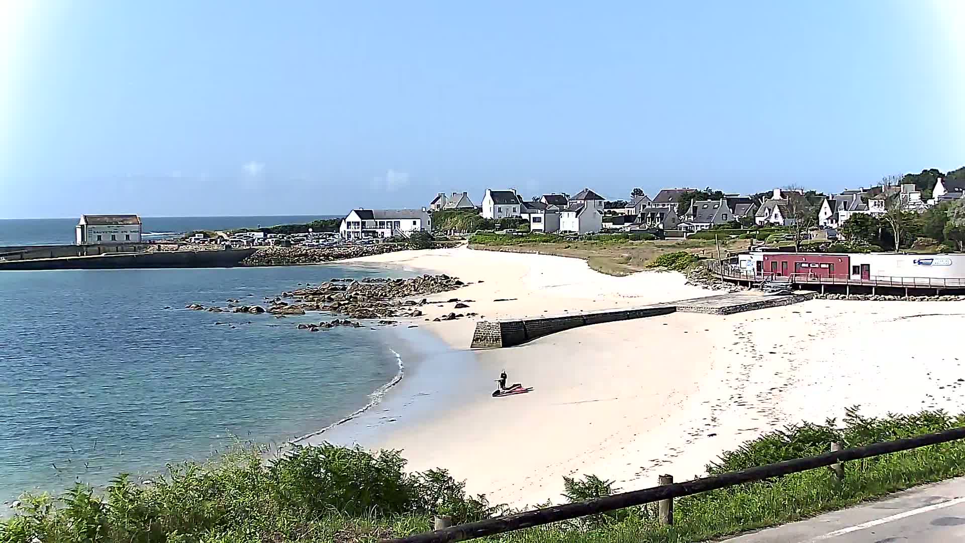 A sandy beach curves around a calm bay, next to a small village of white houses under a clear blue sky.