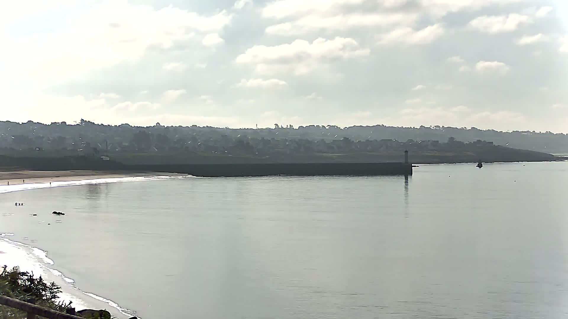 A calm, overcast day reveals a sandy beach curving into a bay, with a distant town nestled behind a low-lying wall and a small lighthouse.