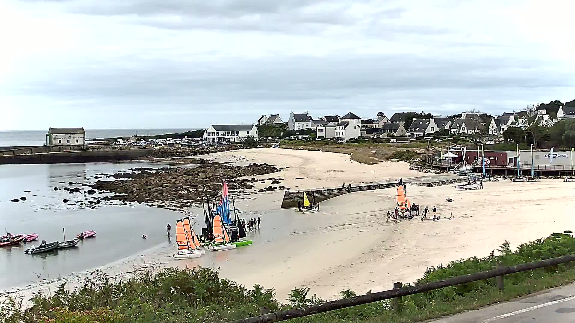 A sandy beach with several sailboats and people on the shore, under an overcast sky, near a small village.