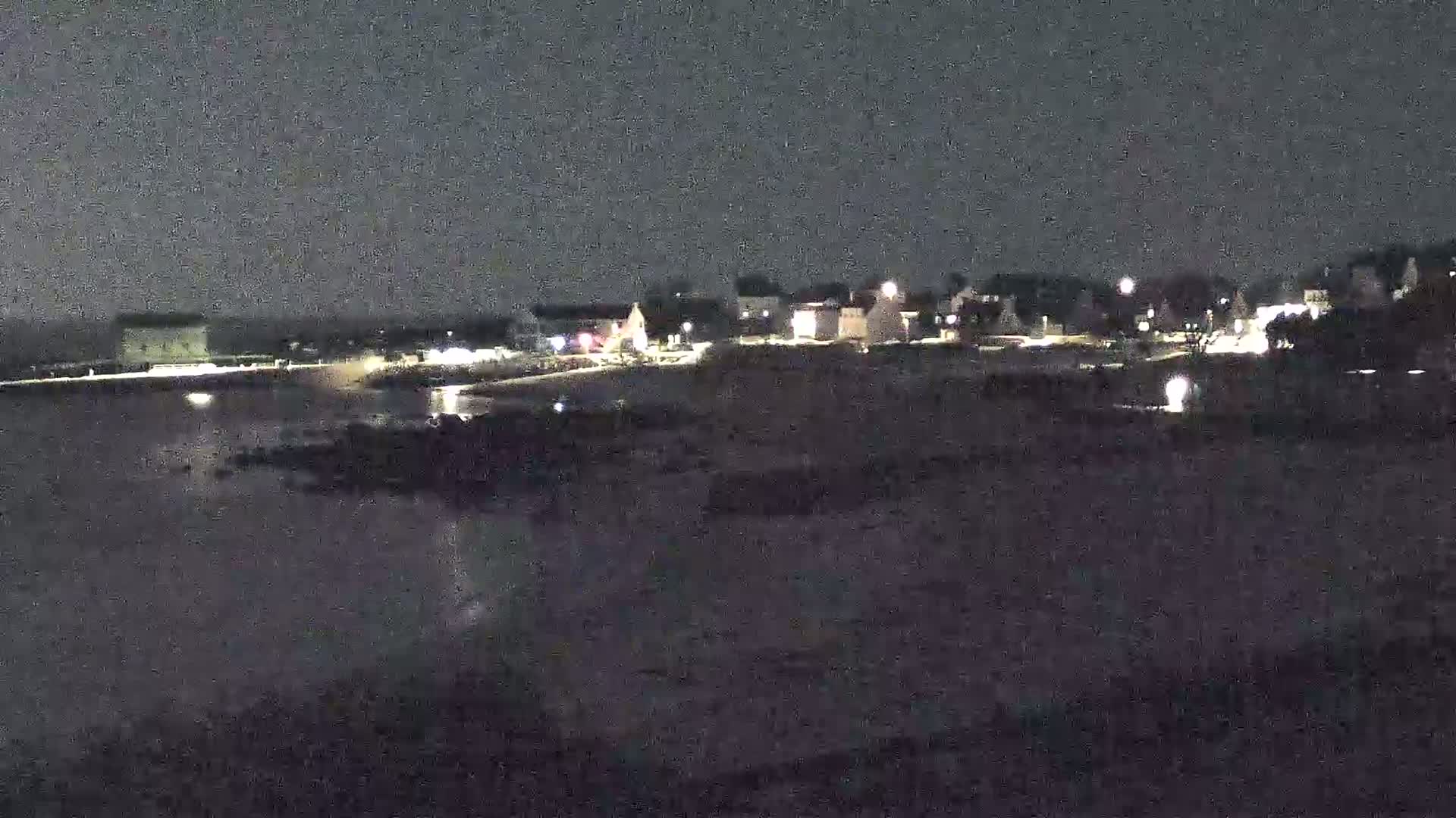 A nighttime view shows a dark body of water reflecting the lights of a small town on the far shore under a dark, overcast sky.