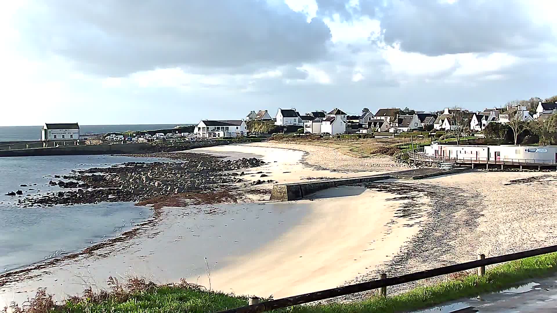 A coastal village featuring white houses overlooks a curved sandy and rocky beach with calm water and various waterfront structures, all under a partly cloudy sky.