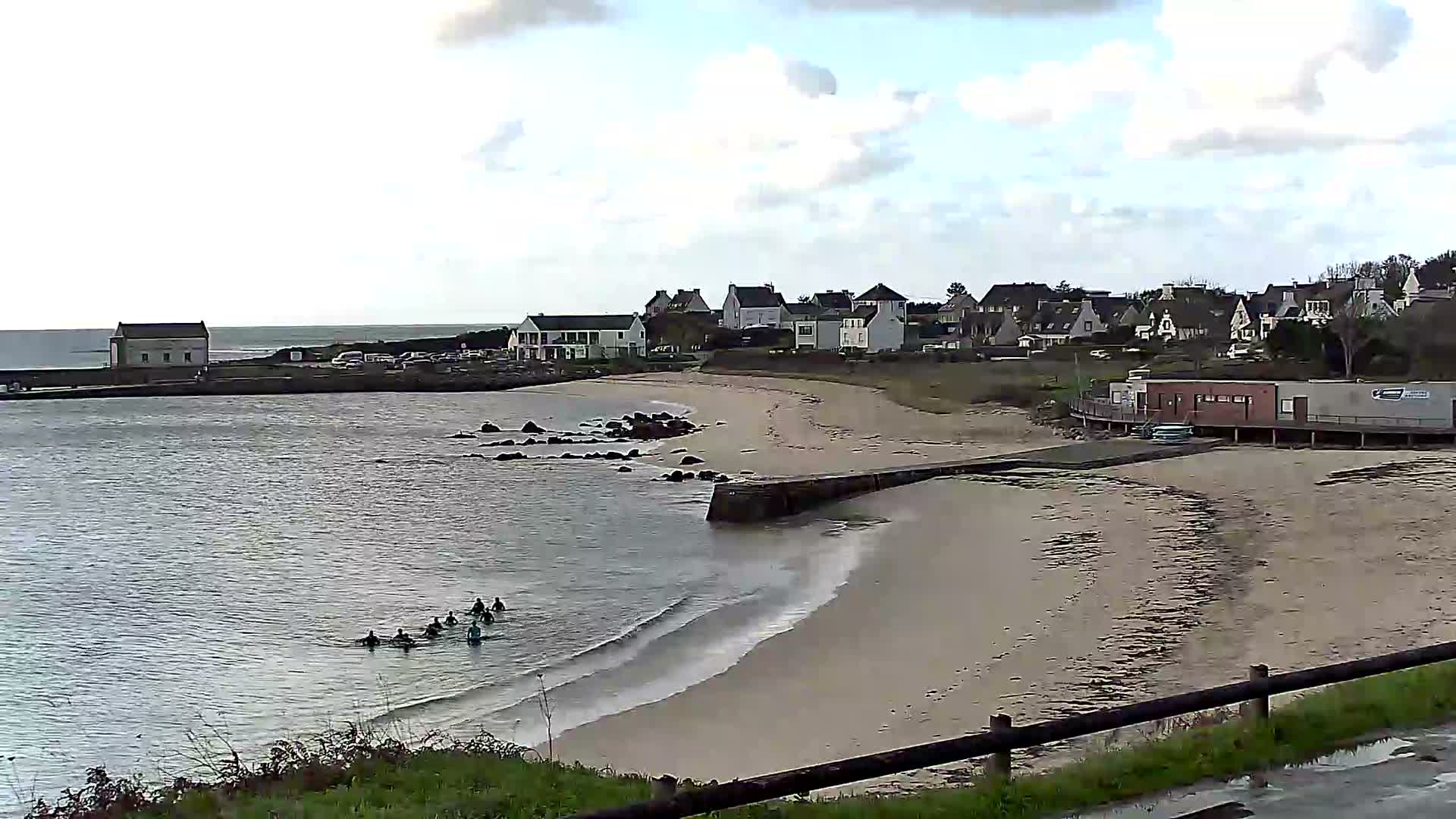 A panoramic view reveals a curving sandy beach with a small group of people in wetsuits in the calm water, bordered by a village of houses nestled on a gentle hillside under a partly cloudy sky.