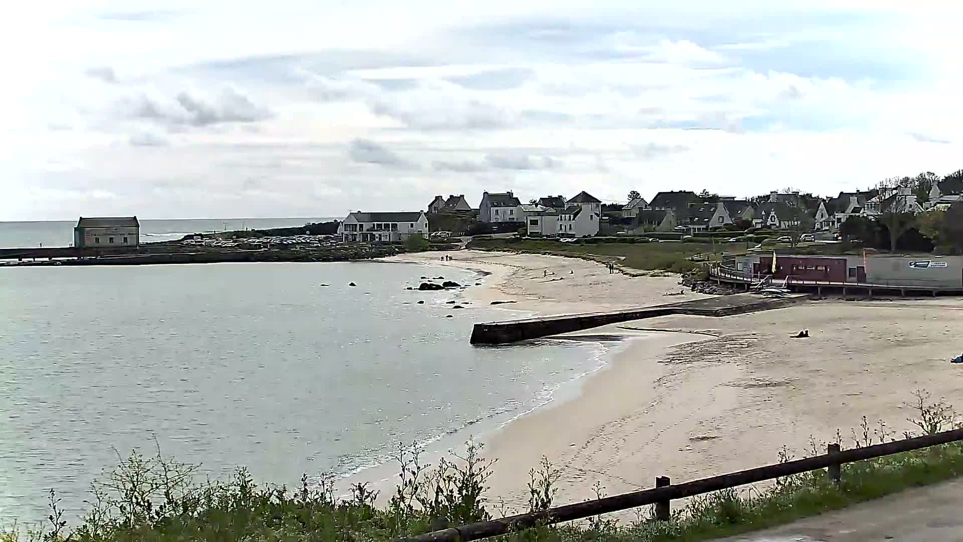 A panoramic view reveals a curving sandy beach with a small group of people in wetsuits in the calm water, bordered by a village of houses nestled on a gentle hillside under a partly cloudy sky.