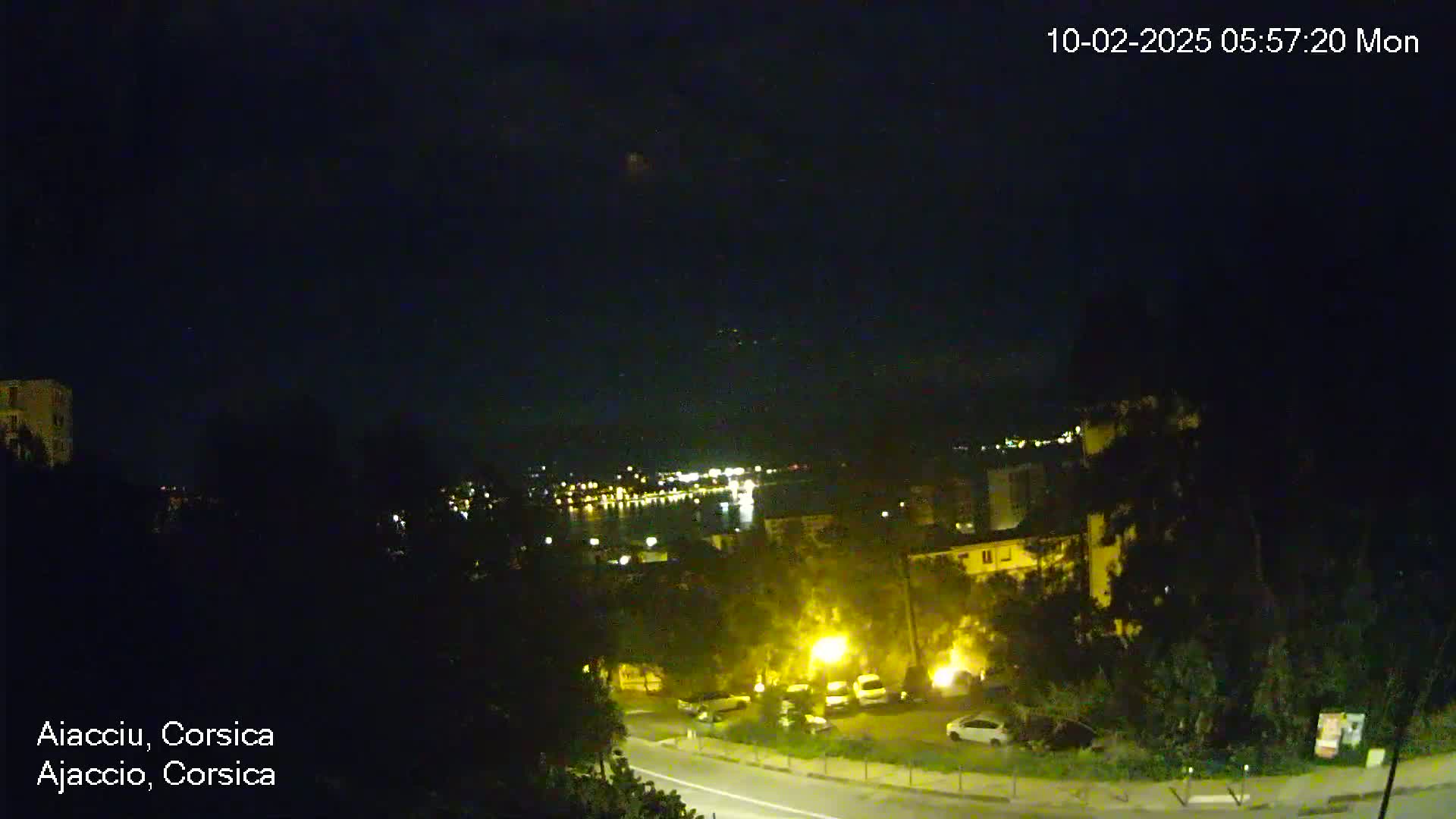 A nighttime, high-angle view shows a coastal town with a road, parked cars, and buildings illuminated by streetlights, overlooking a dark body of water with faintly visible lights across it.