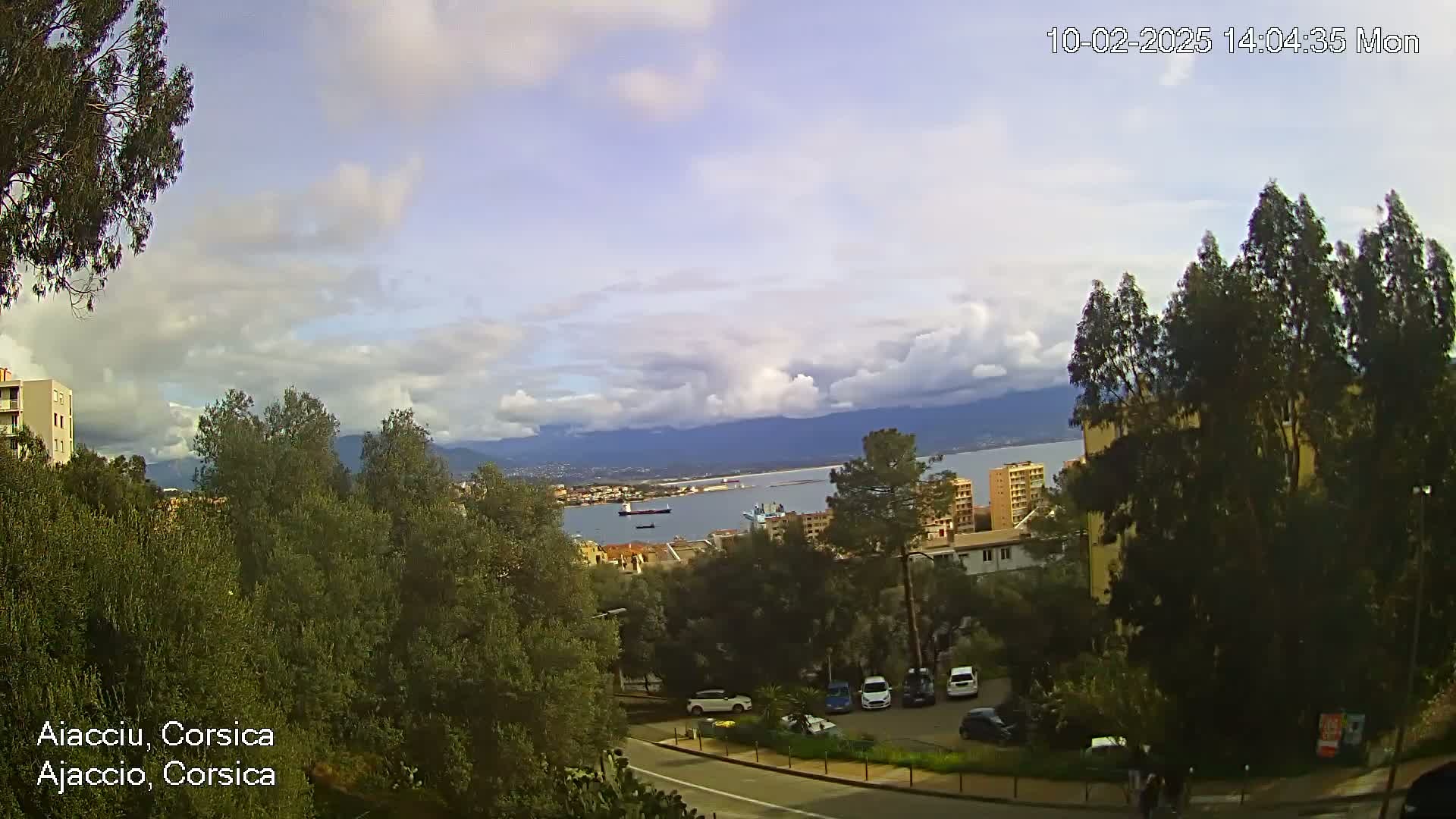 A partly cloudy day overlooks a city with a harbor and mountains in the distance, with several cars parked in a lot and trees in the foreground.