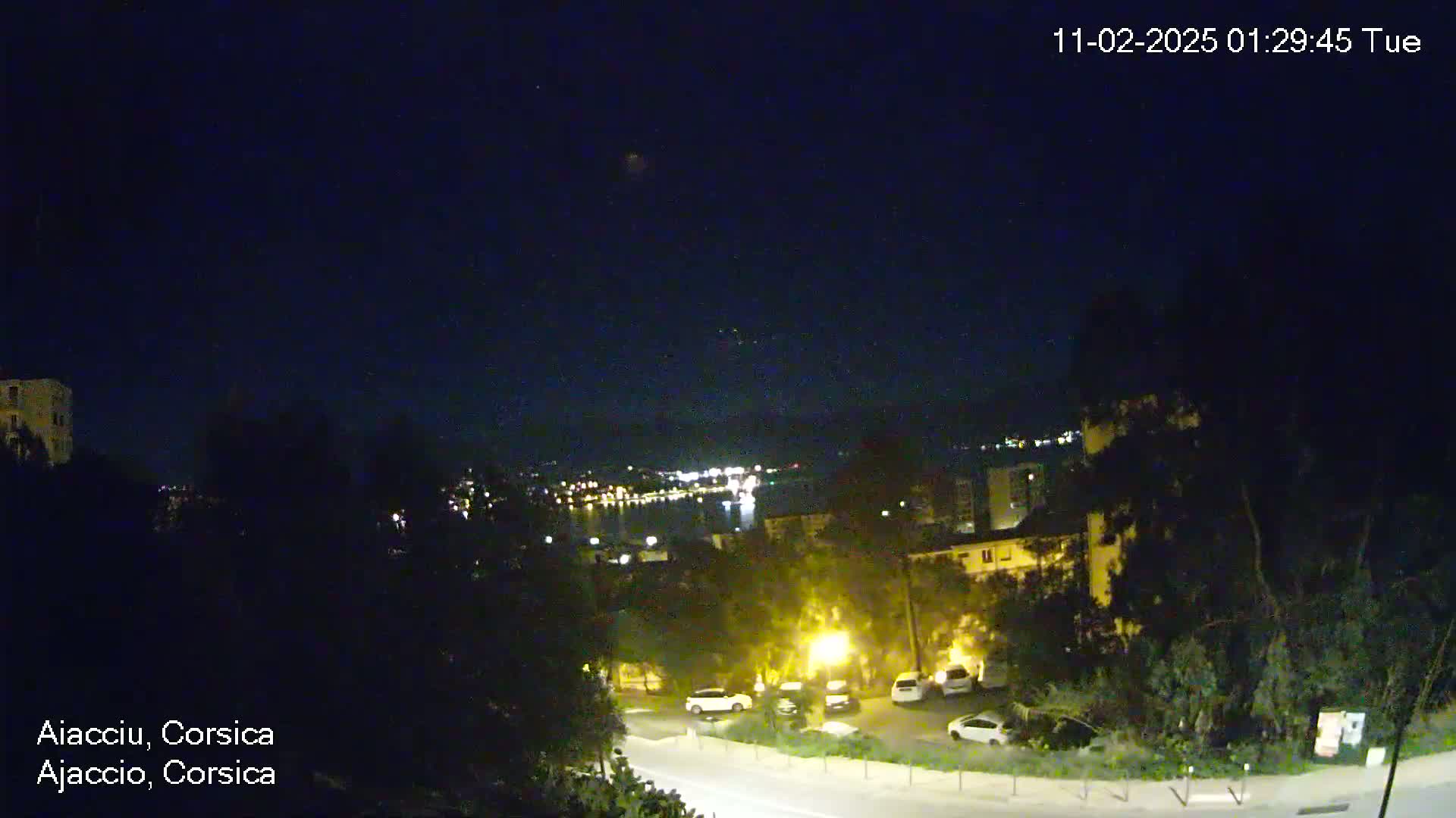 A nighttime, clear-sky view shows a coastal town with parked cars and streetlights, overlooking a dark body of water with distant lights.