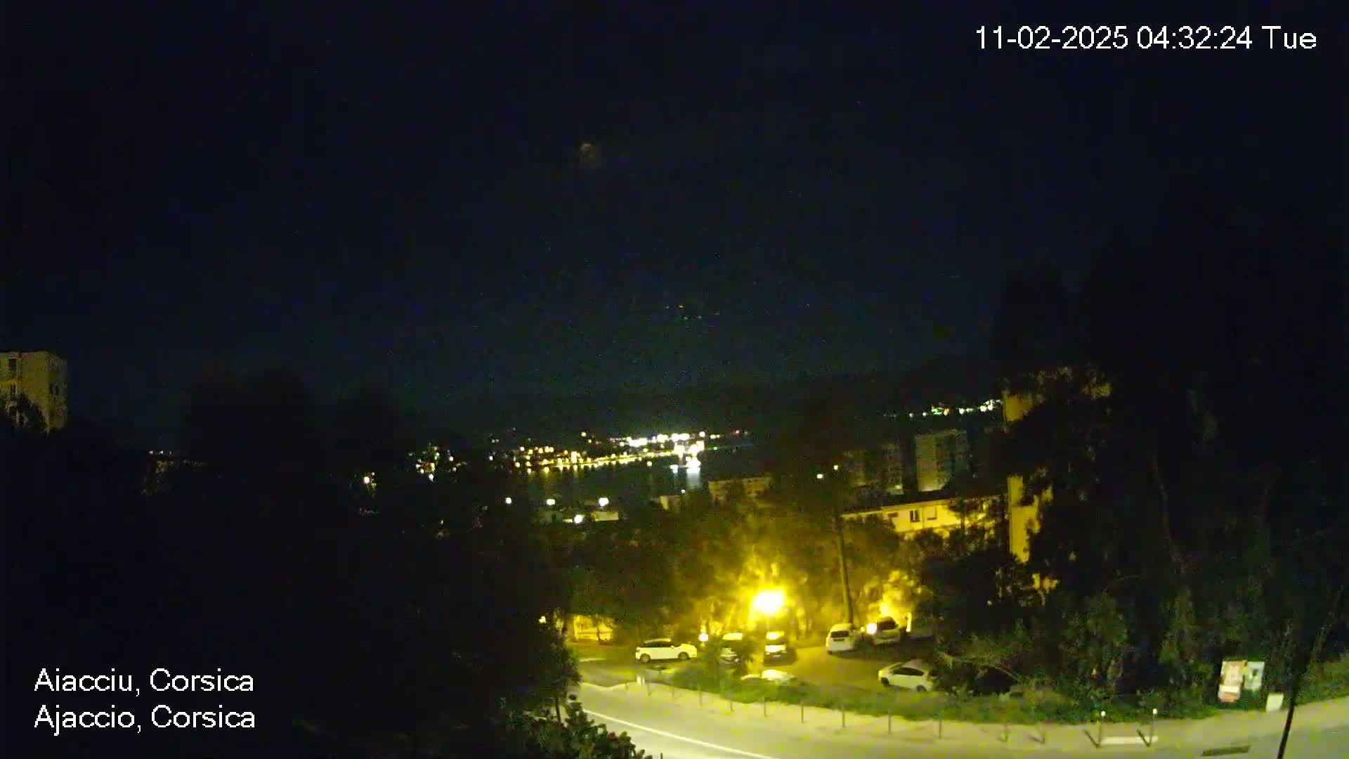 A night-time view of a coastal town, with a clear, dark sky and some cars parked along a road.