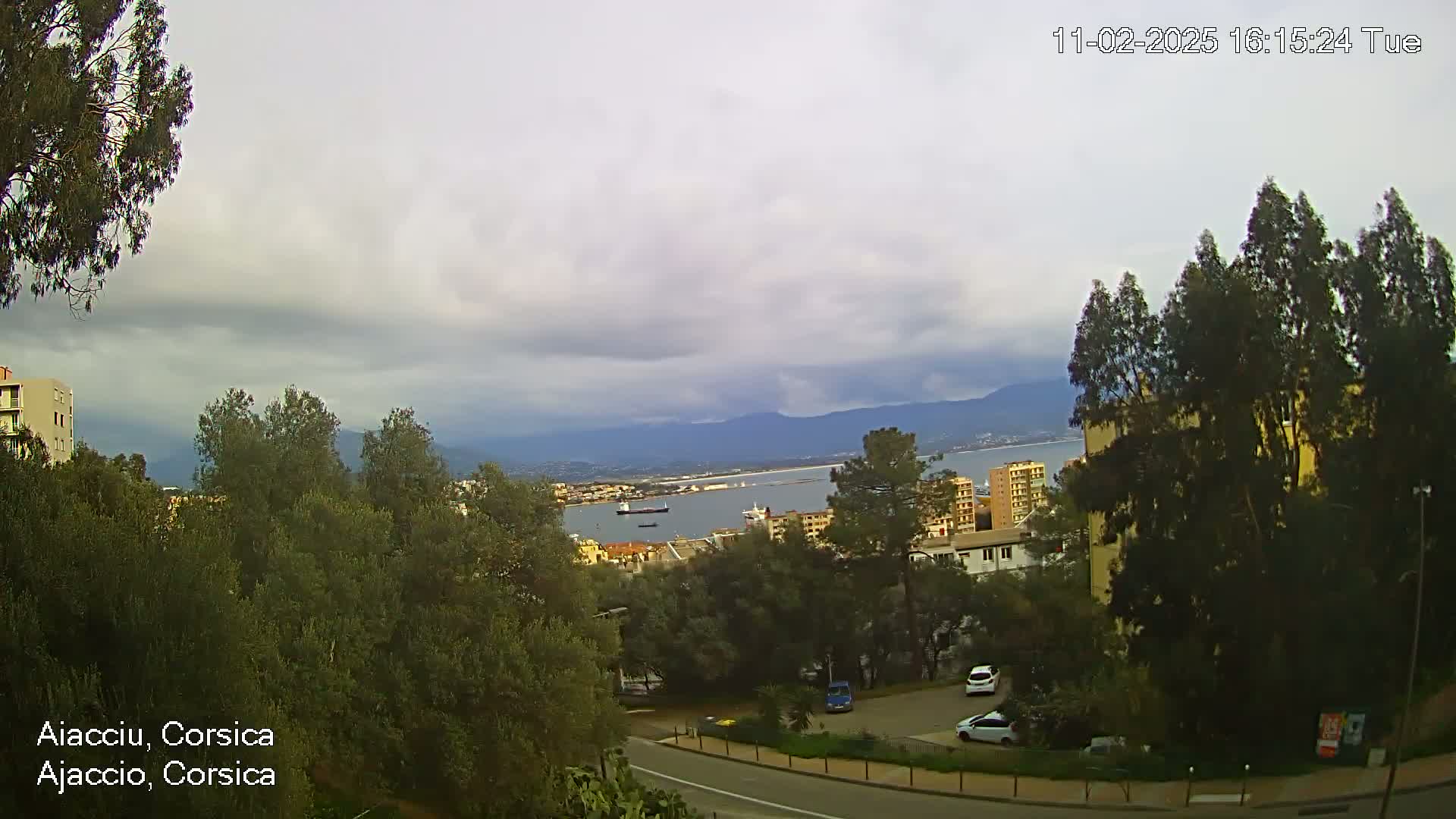 A mostly cloudy day overlooks a coastal city with a harbor, mountains in the distance, and a parking lot with several cars visible.