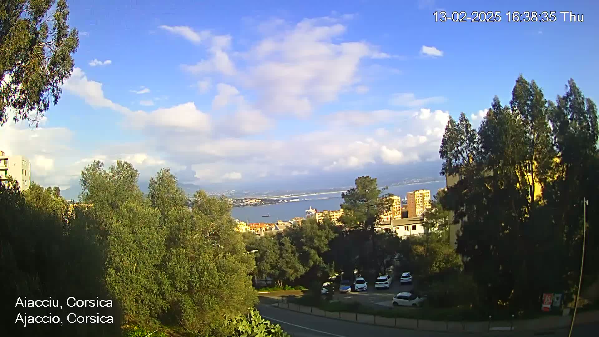 A partly cloudy sky overlooks a coastal city with green trees, buildings, and a parking lot, featuring a body of water in the distance.