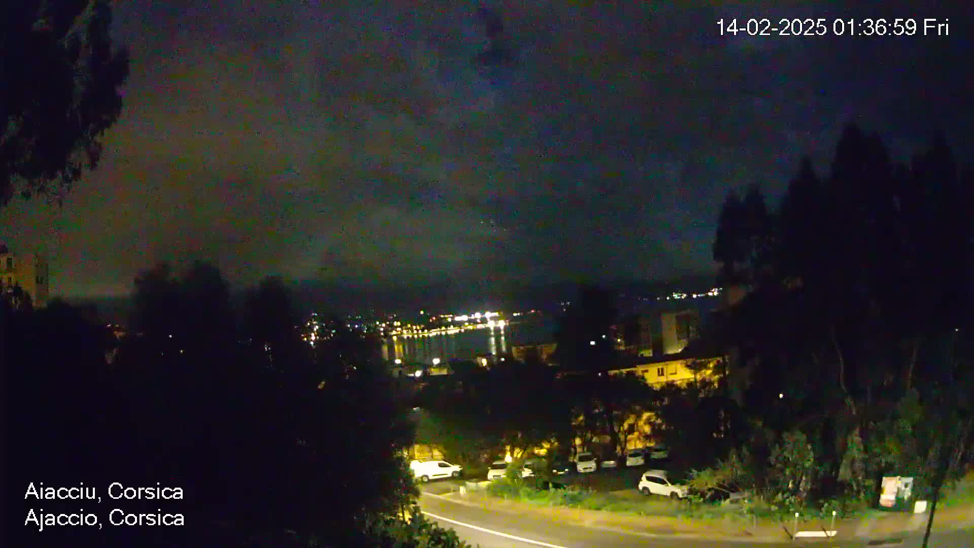 A nighttime view of a city by the water, under a mostly cloudy sky, shows a road with parked cars and buildings illuminated by lights.