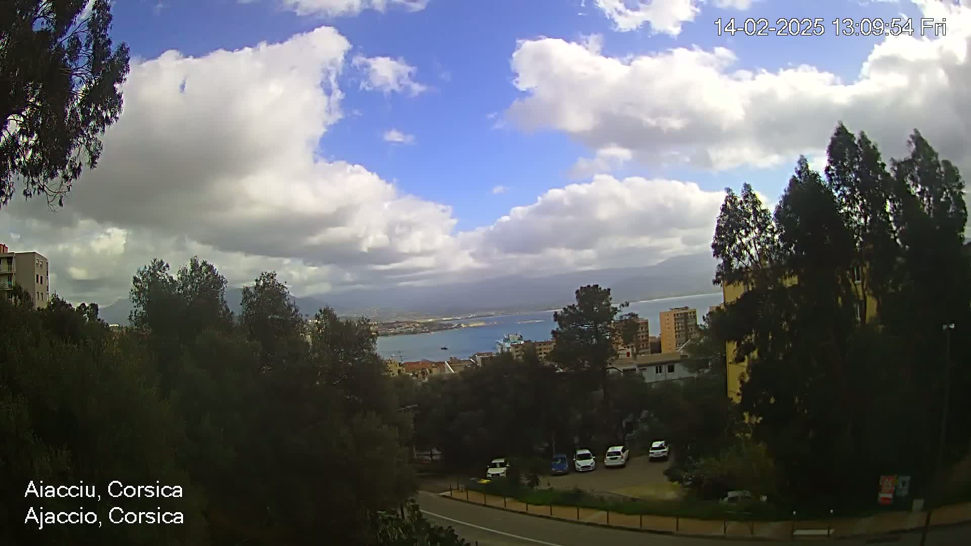 A partly cloudy day reveals a coastal city nestled among green hills and trees, with a body of water visible in the distance.