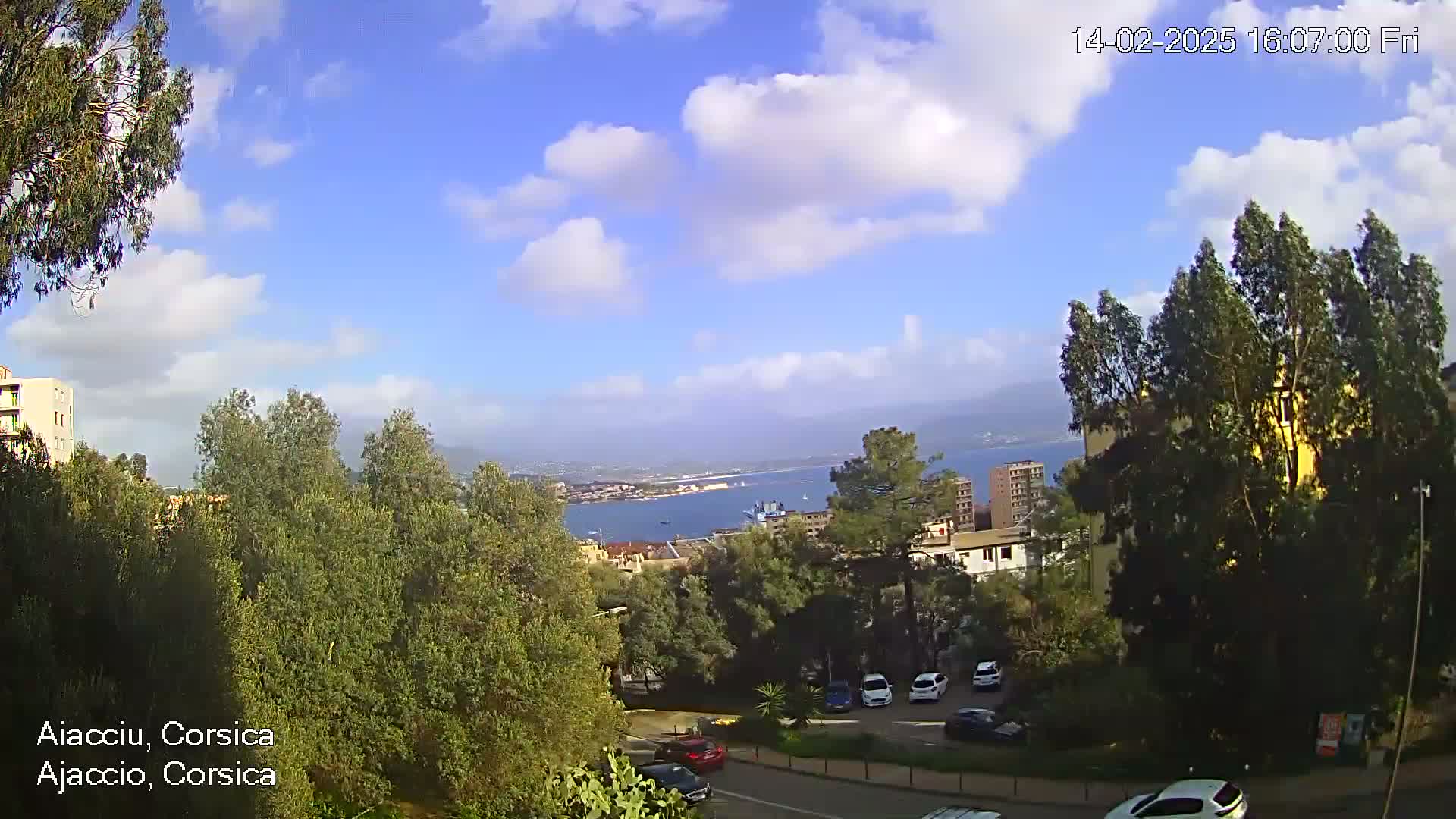 A partly cloudy day overlooks a coastal city with green trees, buildings, and parked cars, a body of water visible in the distance.