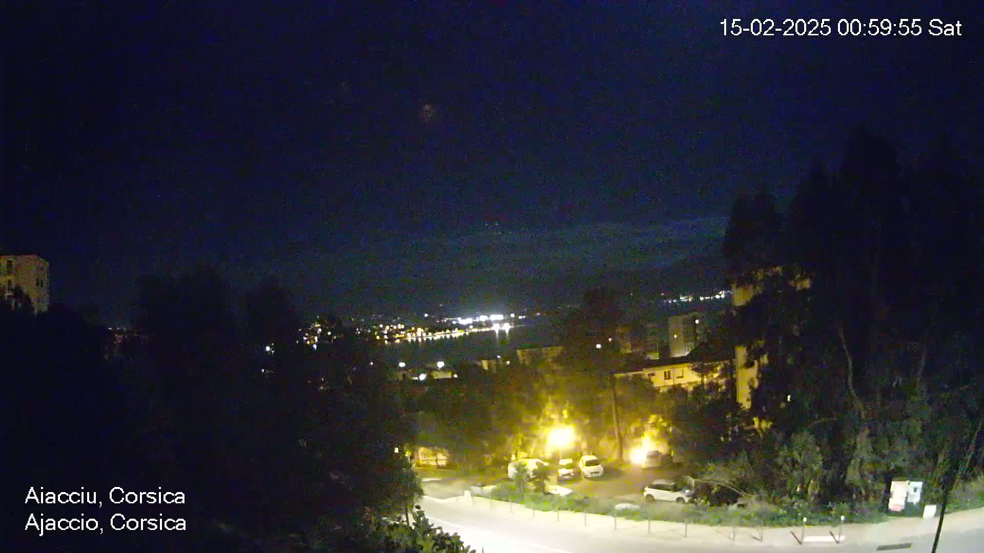 A nighttime, clear-sky view overlooking a city with dimly lit buildings, roadways, and parked cars, situated near a body of water with faint lights on the opposite shore.