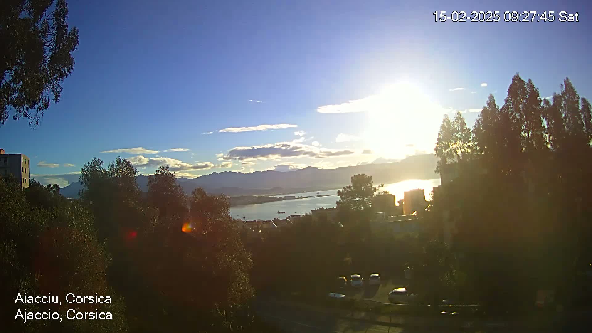 A sunny day with some clouds overlooks a calm body of water, mountains in the distance, and a town nestled amongst trees.