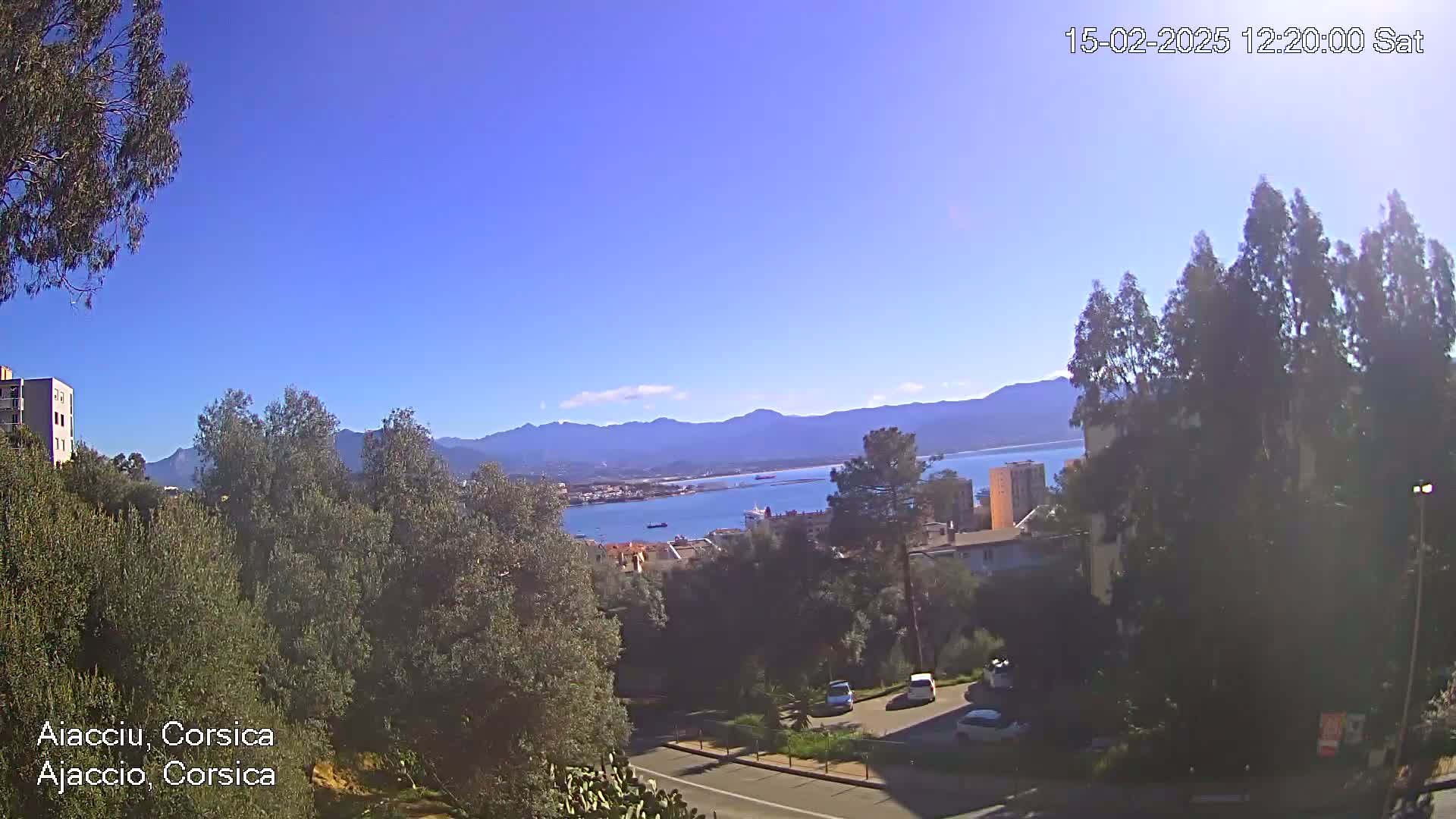 A sunny view of a coastal town nestled amongst green trees and mountains, overlooking a calm body of water.