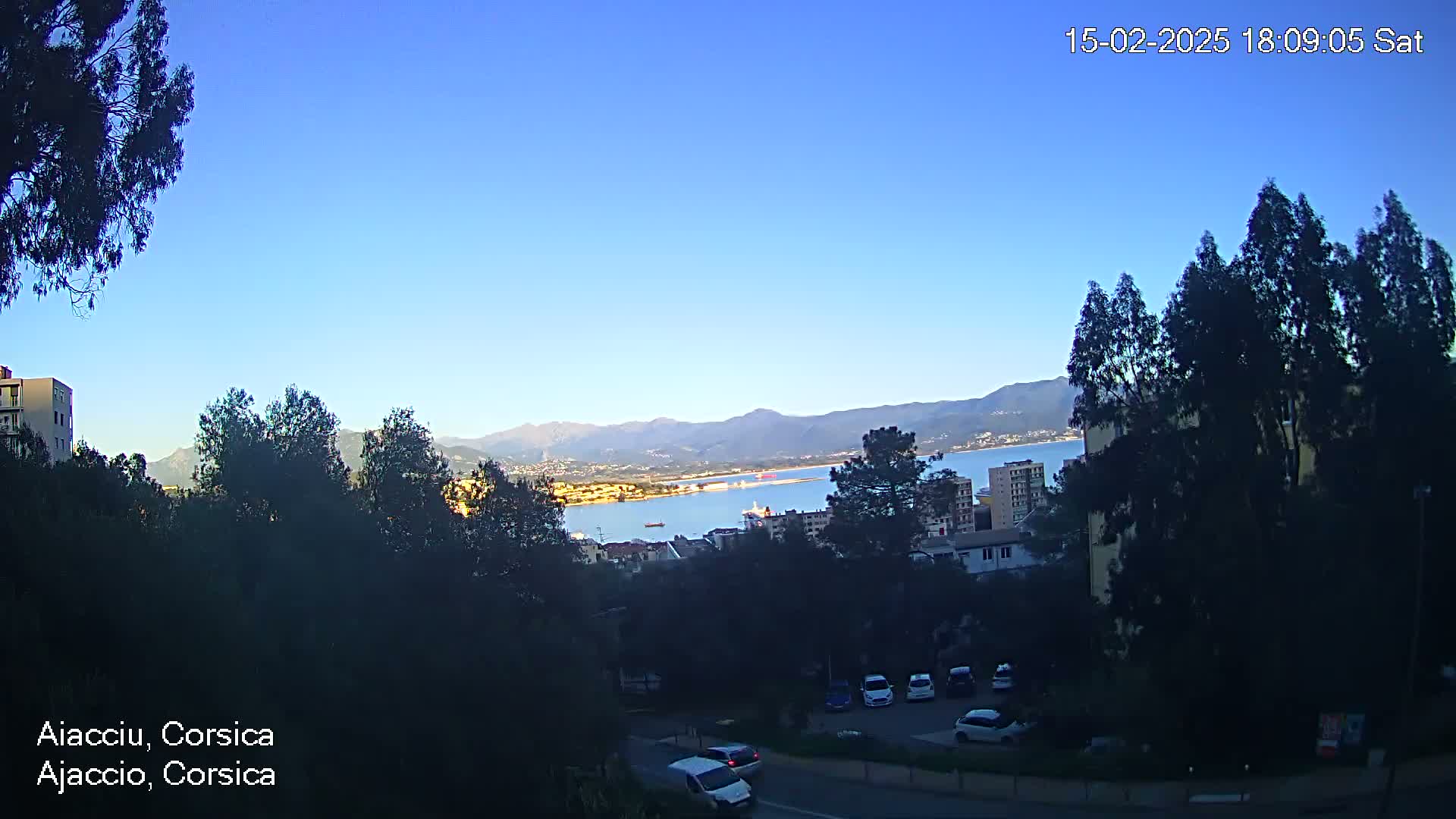 A clear, sunny day reveals a coastal cityscape with a calm body of water, mountains in the distance, and a parking lot with several cars.