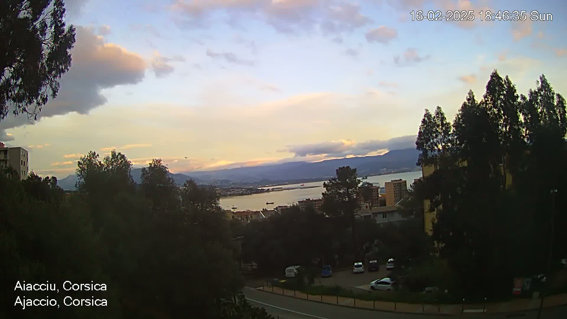 A partly cloudy sunset over a coastal town with a calm body of water, mountains in the distance, and a parking lot in the foreground.