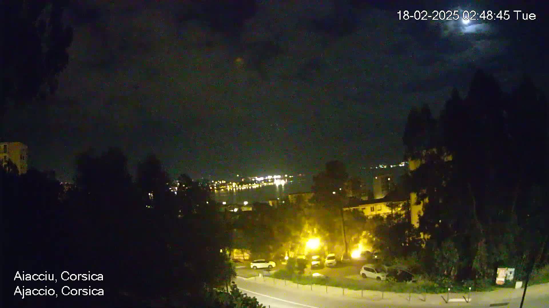 A nighttime, partly cloudy view of a city overlooking a body of water with dimly lit buildings and parked cars.