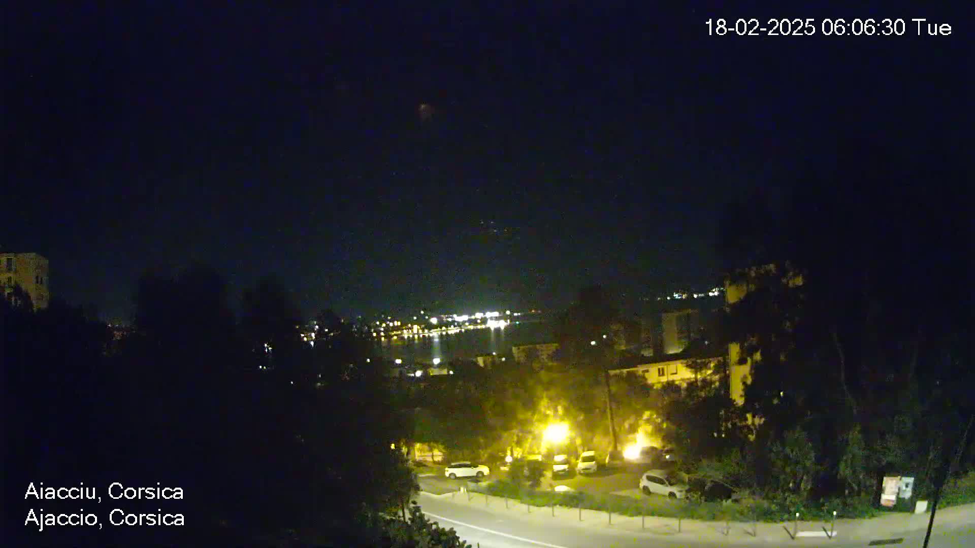 A nighttime view of a town overlooking a body of water with city lights visible in the distance under a clear, dark sky.