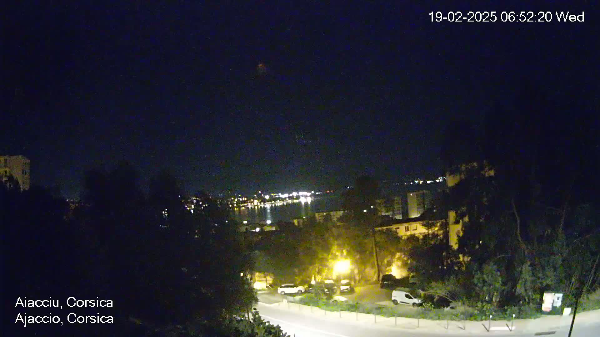 A nighttime, clear-sky view overlooking a city next to a body of water, with cars parked along a road below.