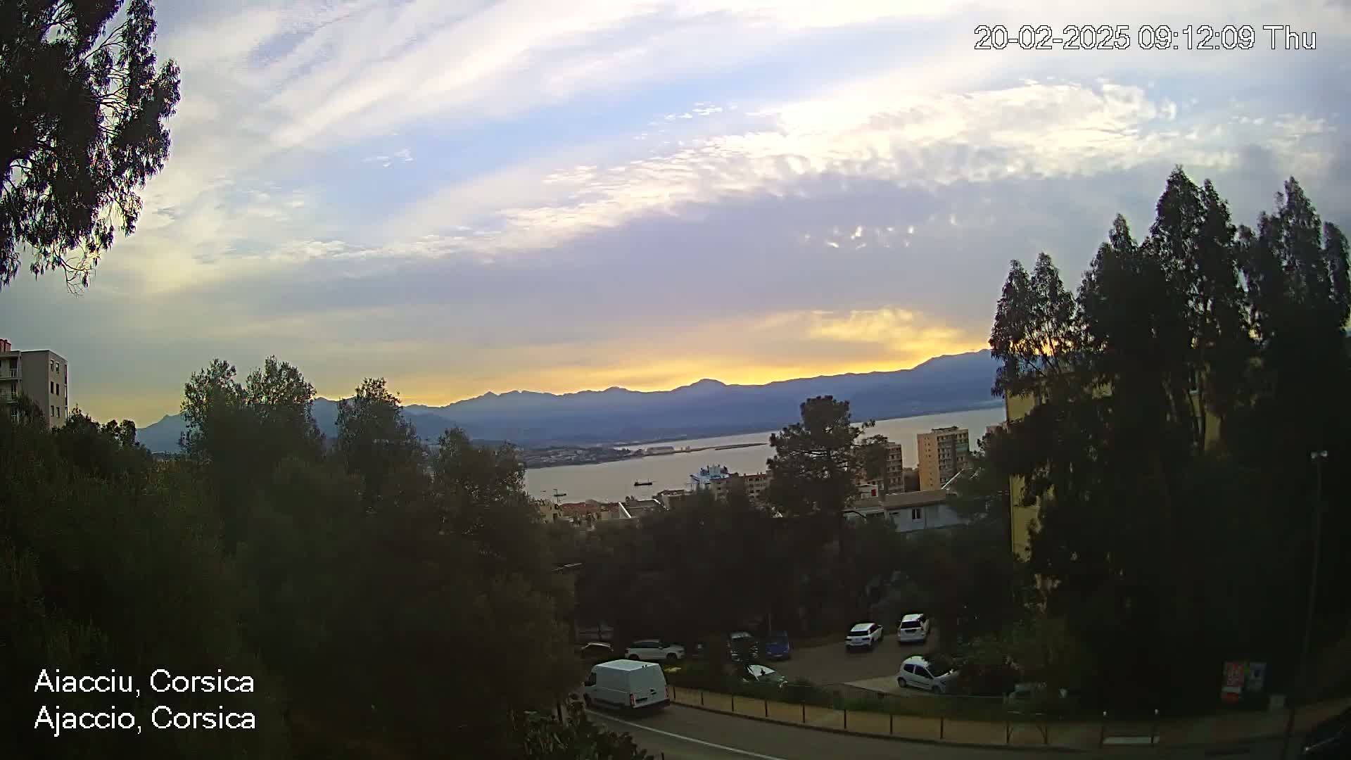 A partly cloudy sunset over a calm body of water, mountains in the distance, and a hillside with buildings and parked cars.