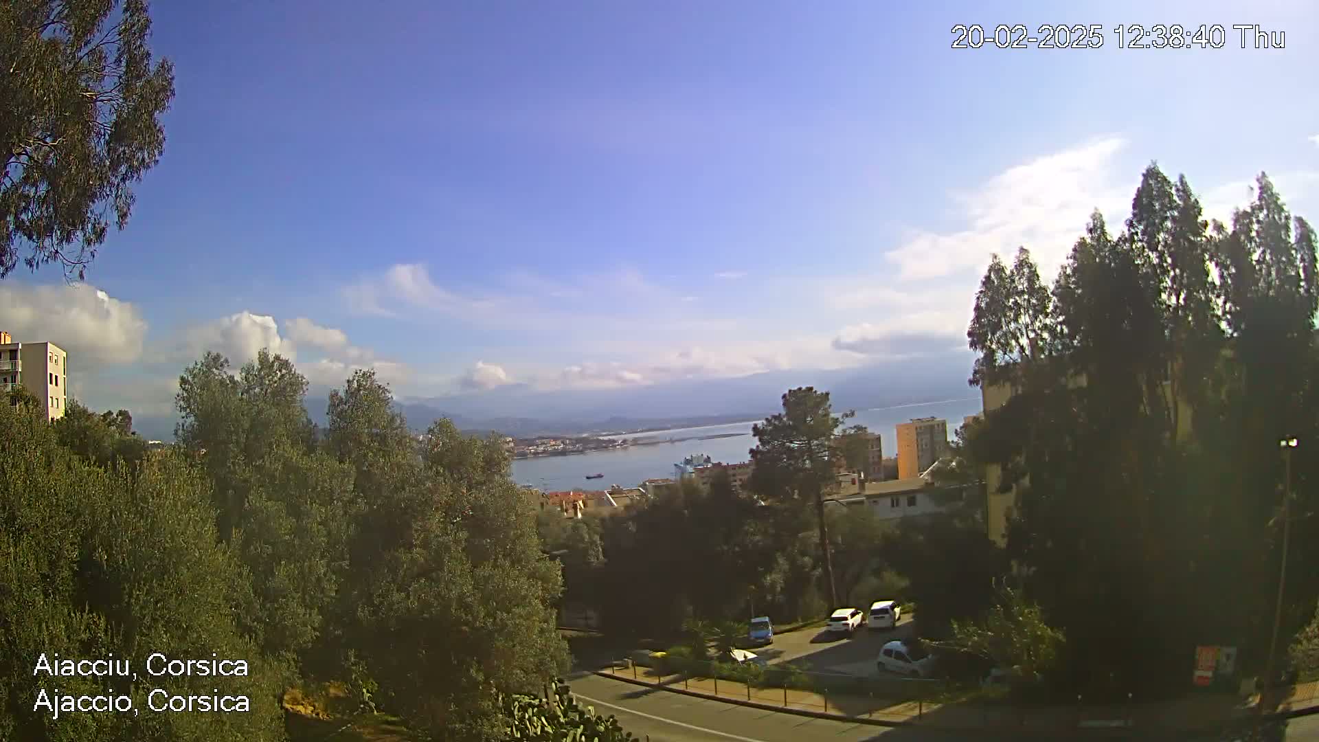 A partly cloudy day reveals a view of a coastal town, with buildings, trees, parked cars, and a body of water extending to distant mountains.