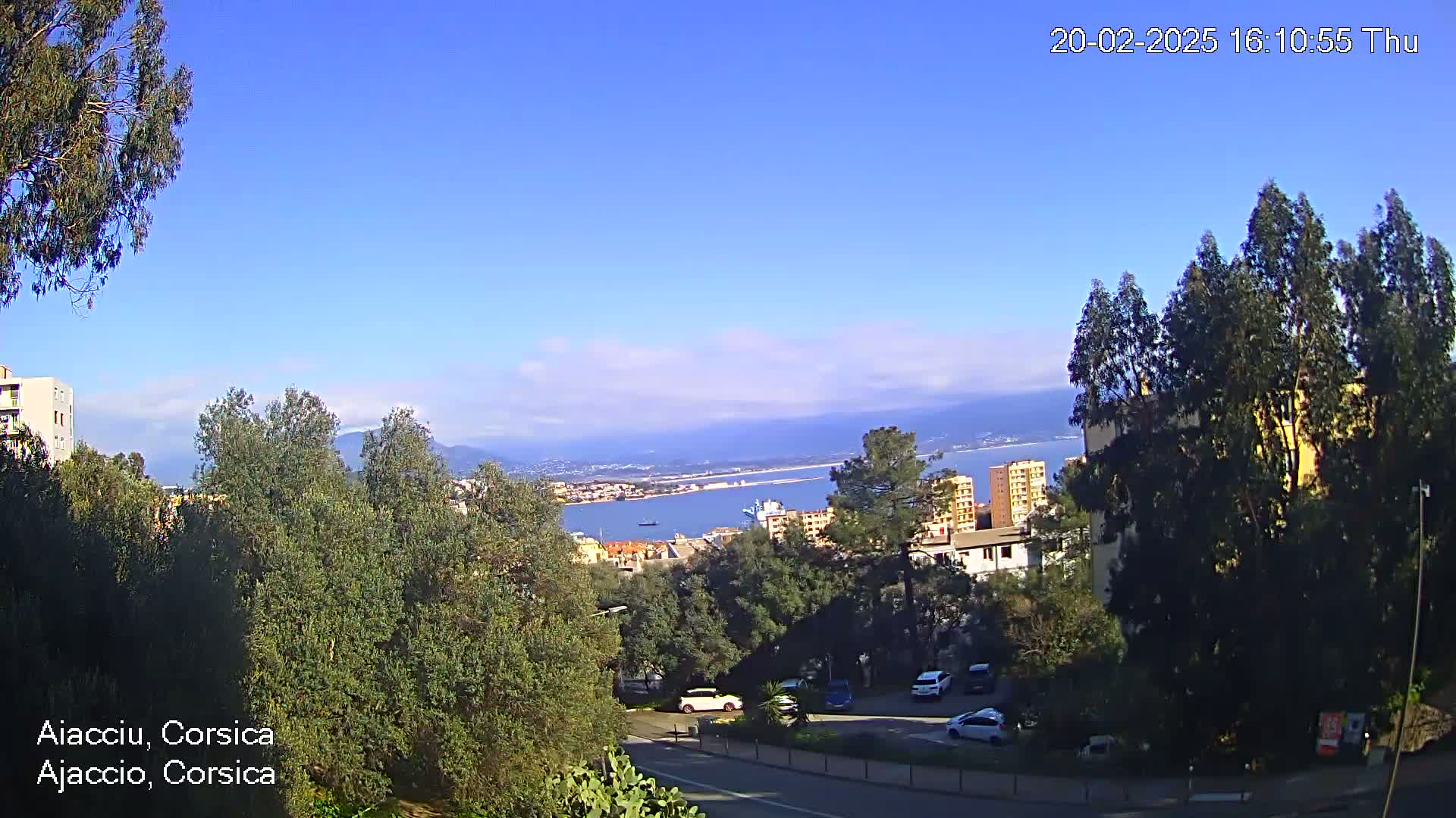 A partly cloudy day reveals a coastal city scene with green trees, buildings, parked cars, and a body of water stretching to distant mountains.