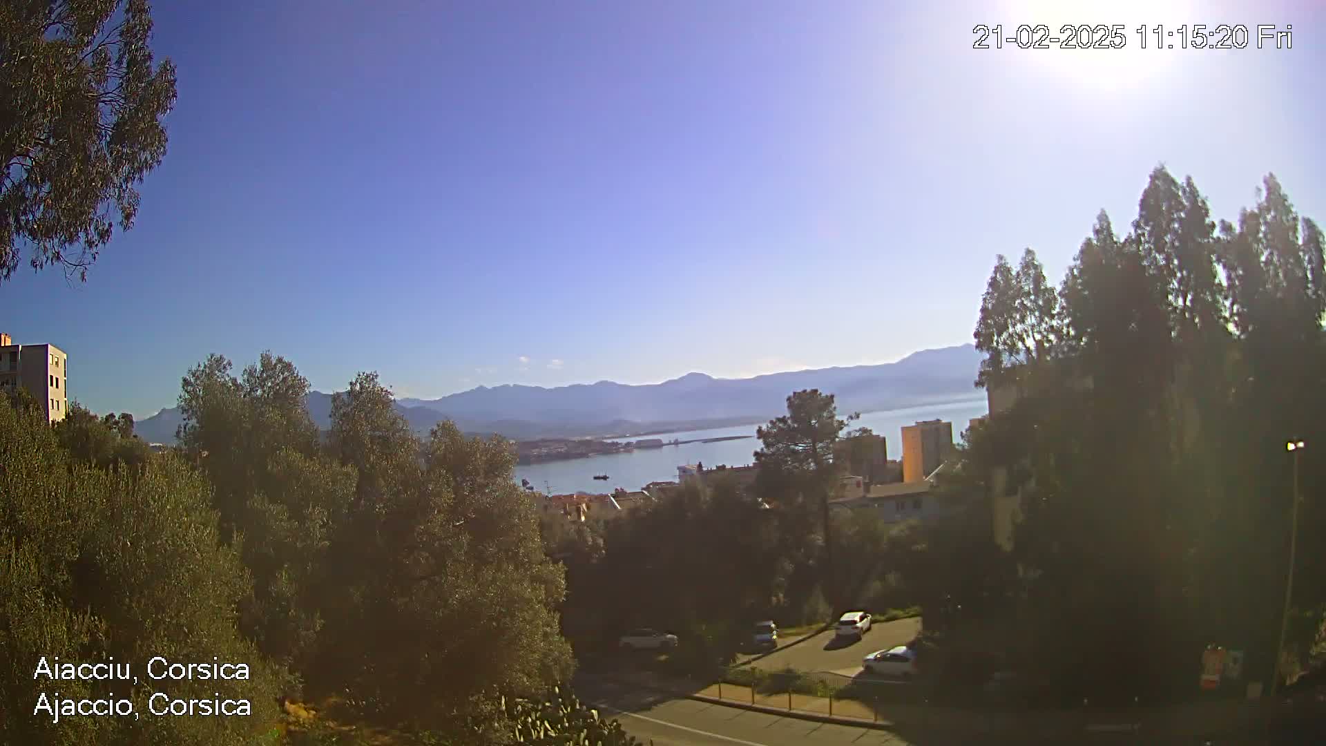 A sunny, clear day overlooks a coastal town nestled between mountains and a calm body of water, with a few cars visible in a parking lot below.