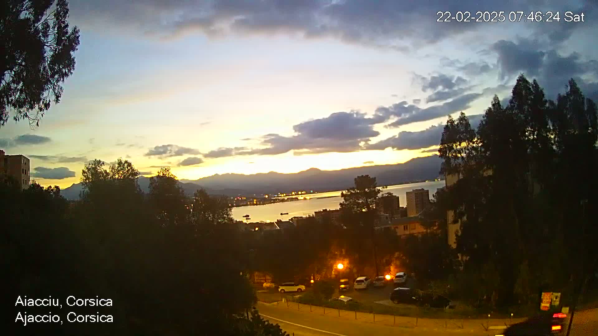A twilight view of a coastal city with buildings, cars, and trees, overlooking a calm body of water and mountains under a partly cloudy sky.
