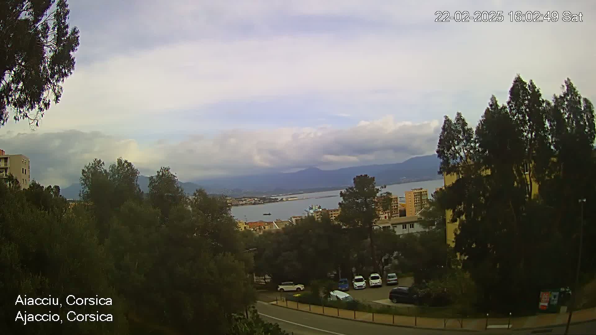 A partly cloudy day overlooks a coastal city with a bay and mountains in the distance, seen from a hilltop vantage point.