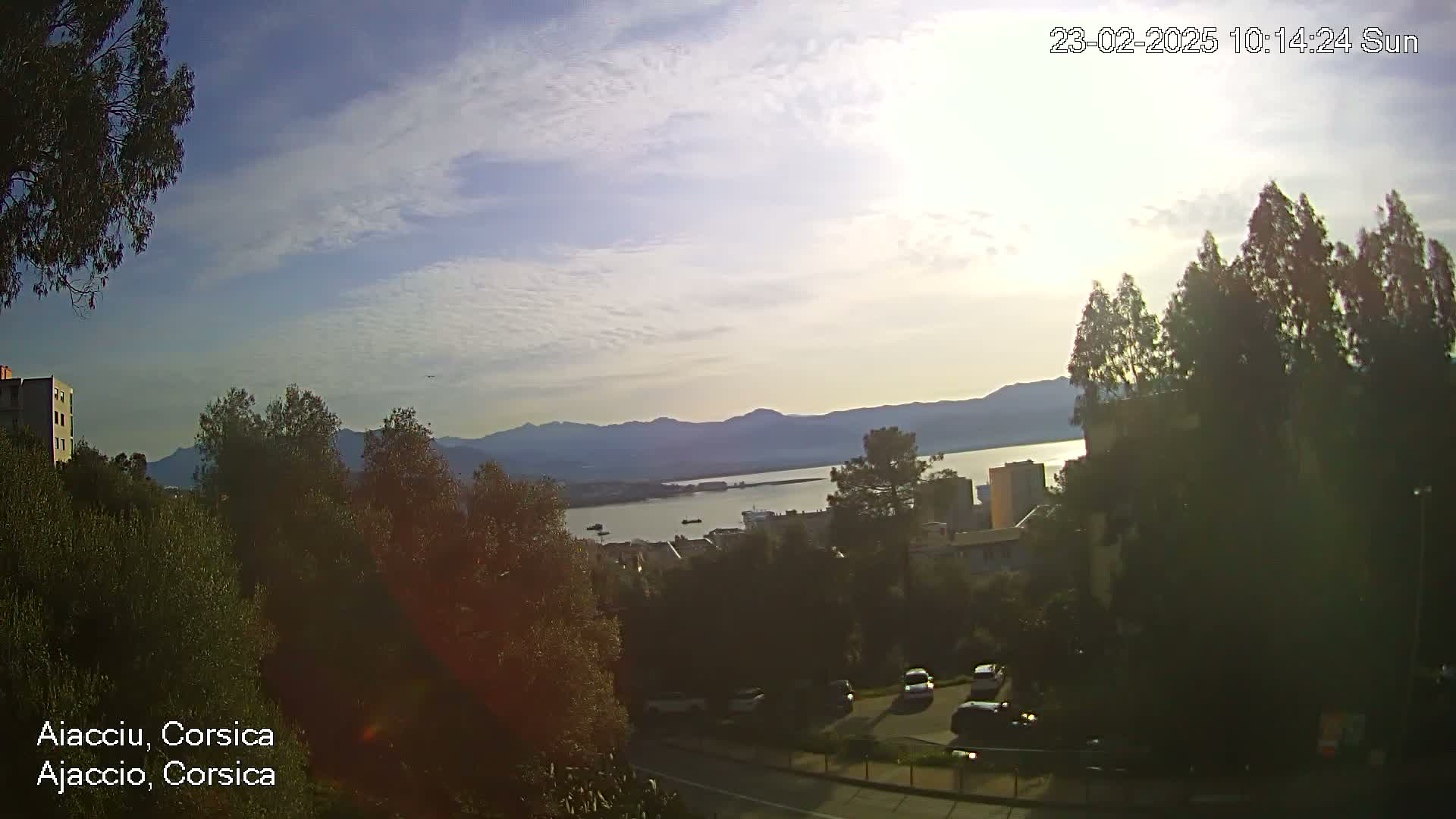 A partly sunny day overlooks a calm bay with mountains in the distance, and several cars parked along a road surrounded by trees and buildings.