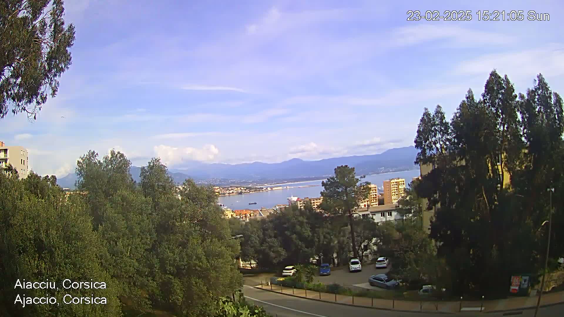 A partly cloudy, sunny day overlooks a coastal town with a harbor, mountains in the distance, and a parking lot nestled among green trees.