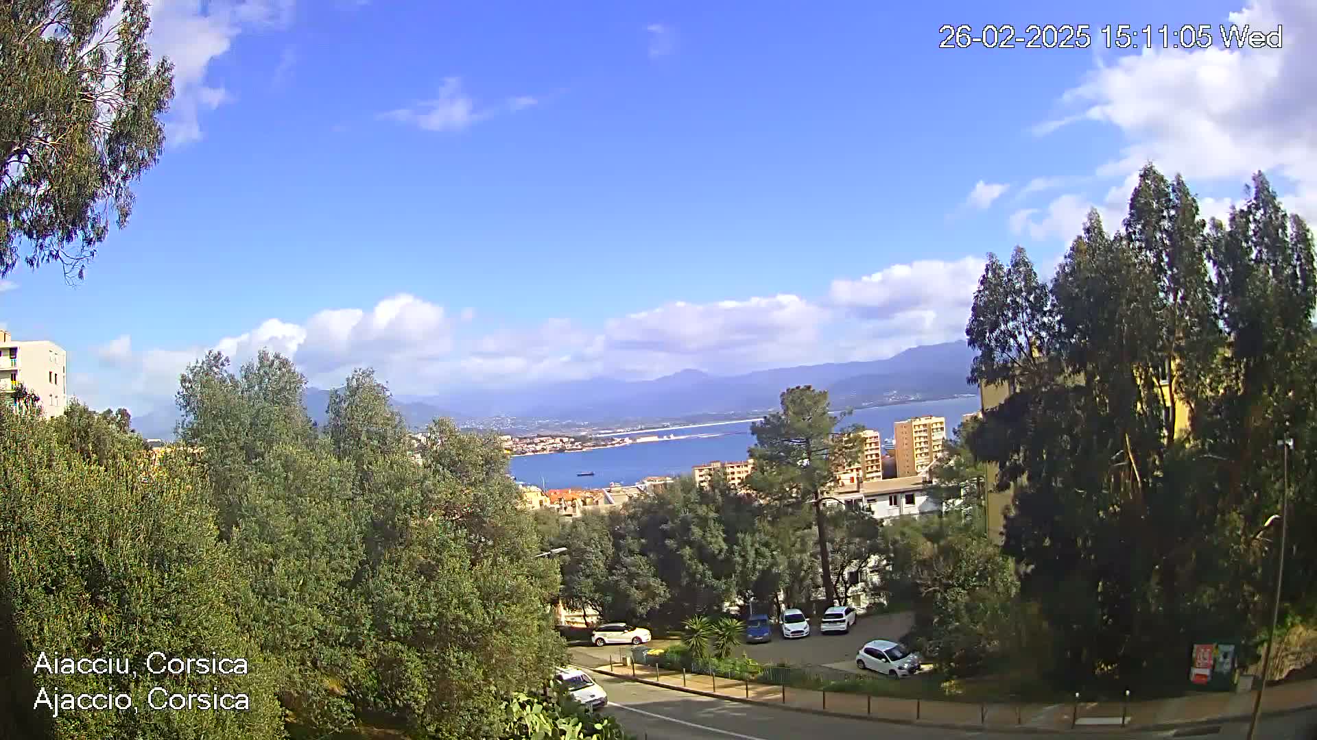 A partly sunny day reveals a coastal town nestled among green trees, overlooking a calm bay and distant mountains.