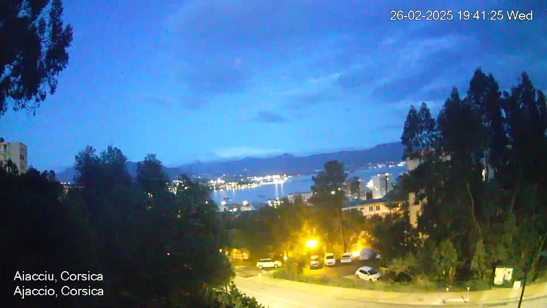 A nighttime view of a city situated on a hill overlooking a calm body of water under a twilight sky with some clouds.