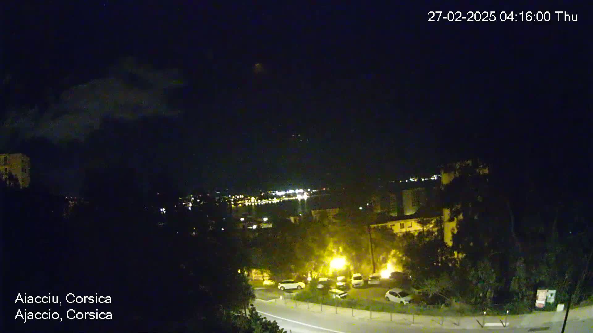 A nighttime, slightly cloudy view of a city by a body of water, with dimly lit buildings and parked cars visible below.
