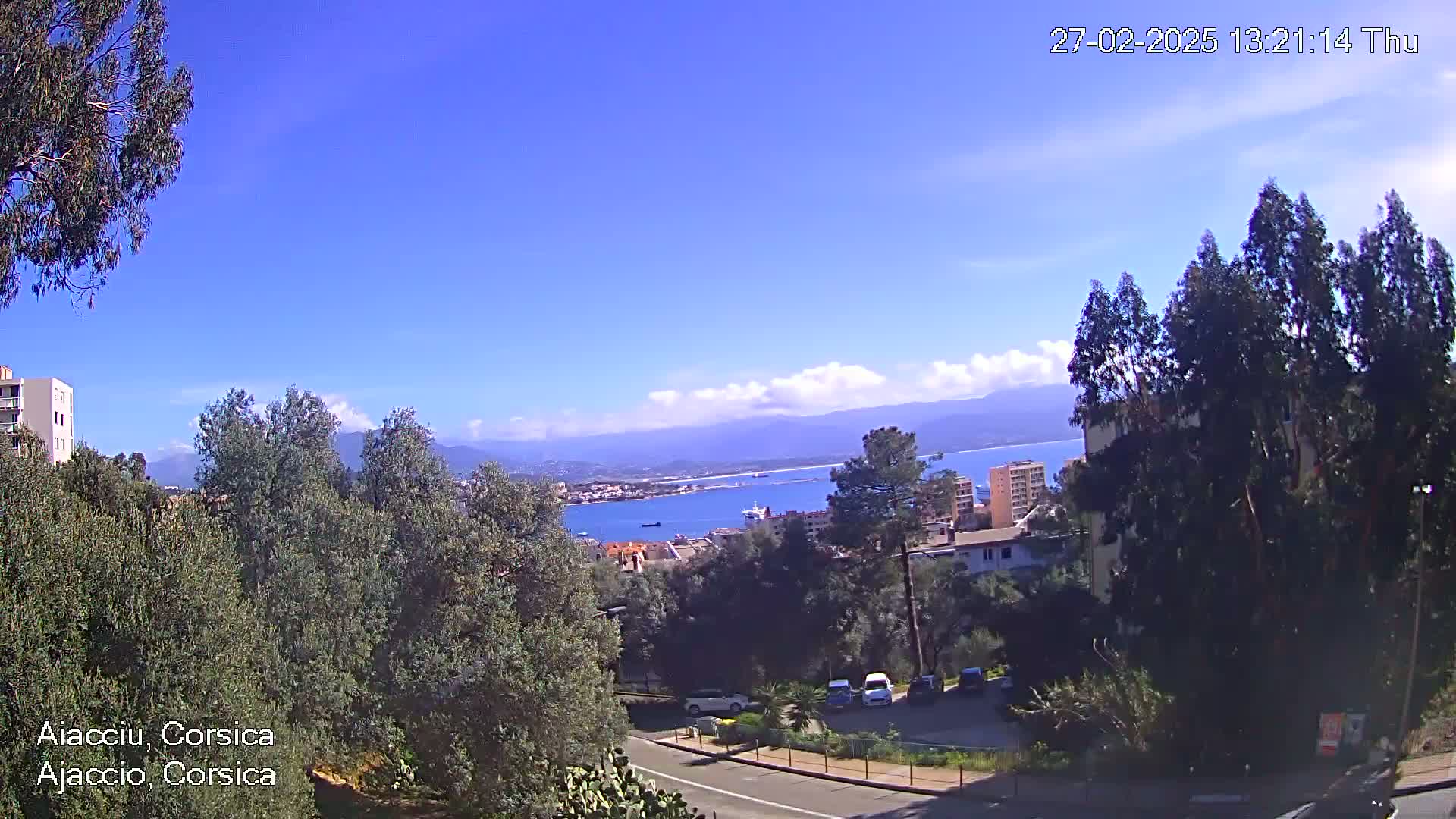 A sunny day overlooks a coastal city with a calm bay, mountains in the distance, and lush green vegetation surrounding a road.