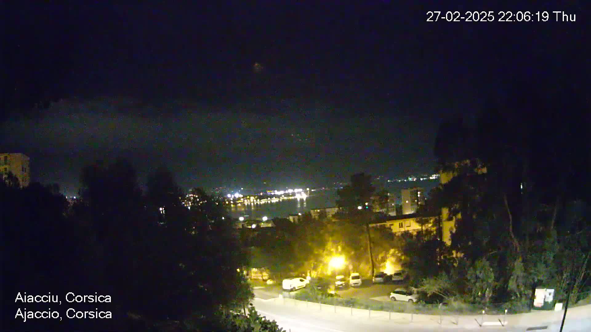 A nighttime view of a town overlooking a body of water, with city lights visible in the distance under a clear, dark sky.