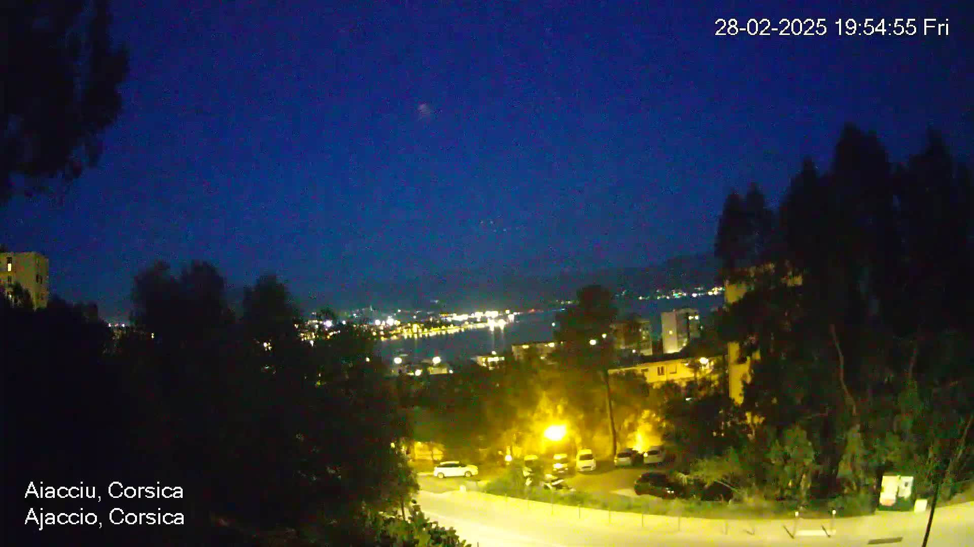 A nighttime view of a city overlooking a body of water with lights visible in the distance under a clear, dark blue sky.