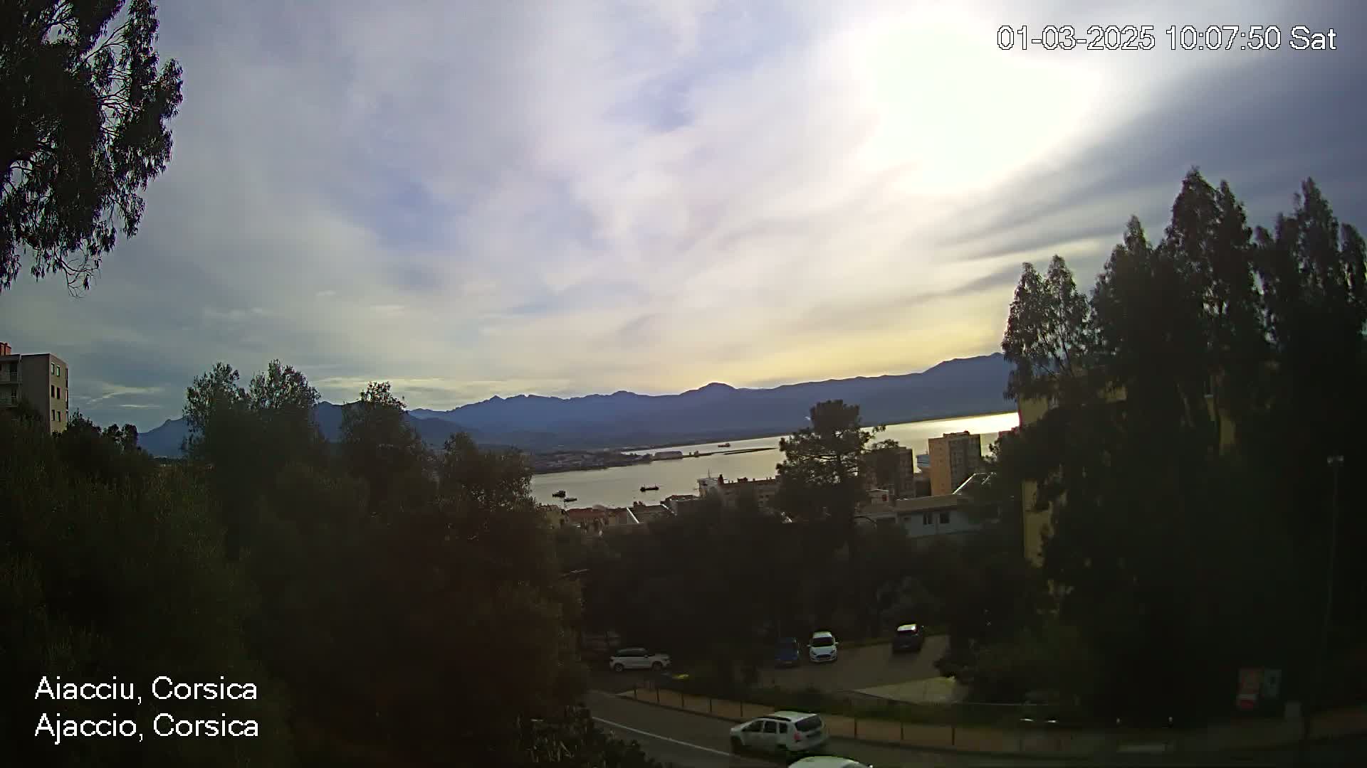 A partly cloudy sunset over a calm body of water, with mountains in the distance and cars parked along a road in the foreground.
