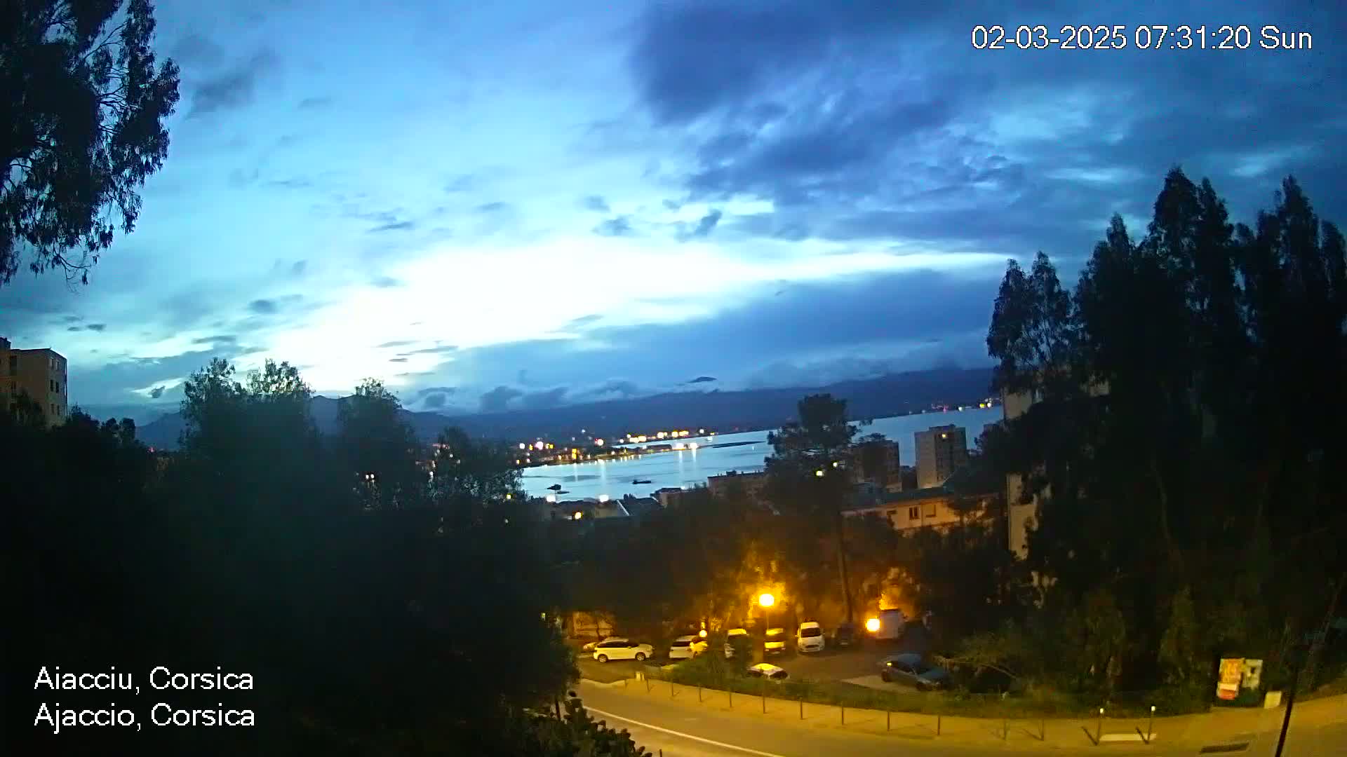 A nighttime view of a city overlooking a calm body of water under a mostly cloudy, blue sky.
