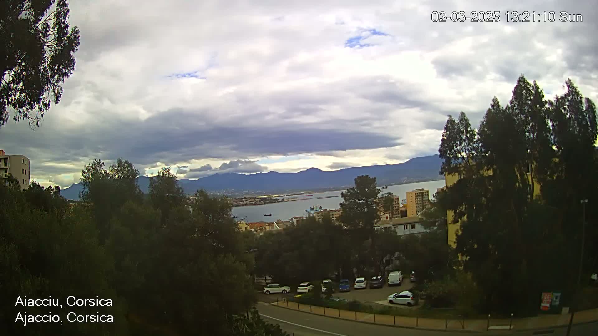 A cloudy, but sunny, view of a coastal city nestled amongst green hills and mountains, with a body of water and parked cars visible.