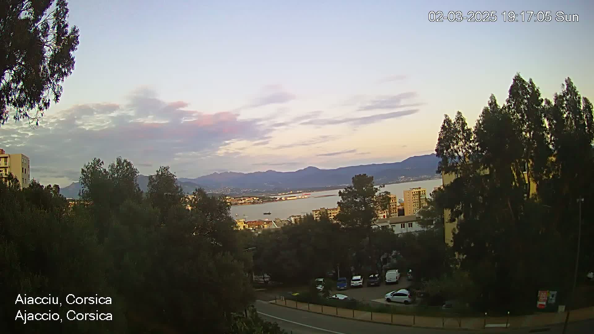A partly cloudy sunset over a body of water with distant mountains, viewed from an elevated position among trees and buildings with a parking lot visible below.